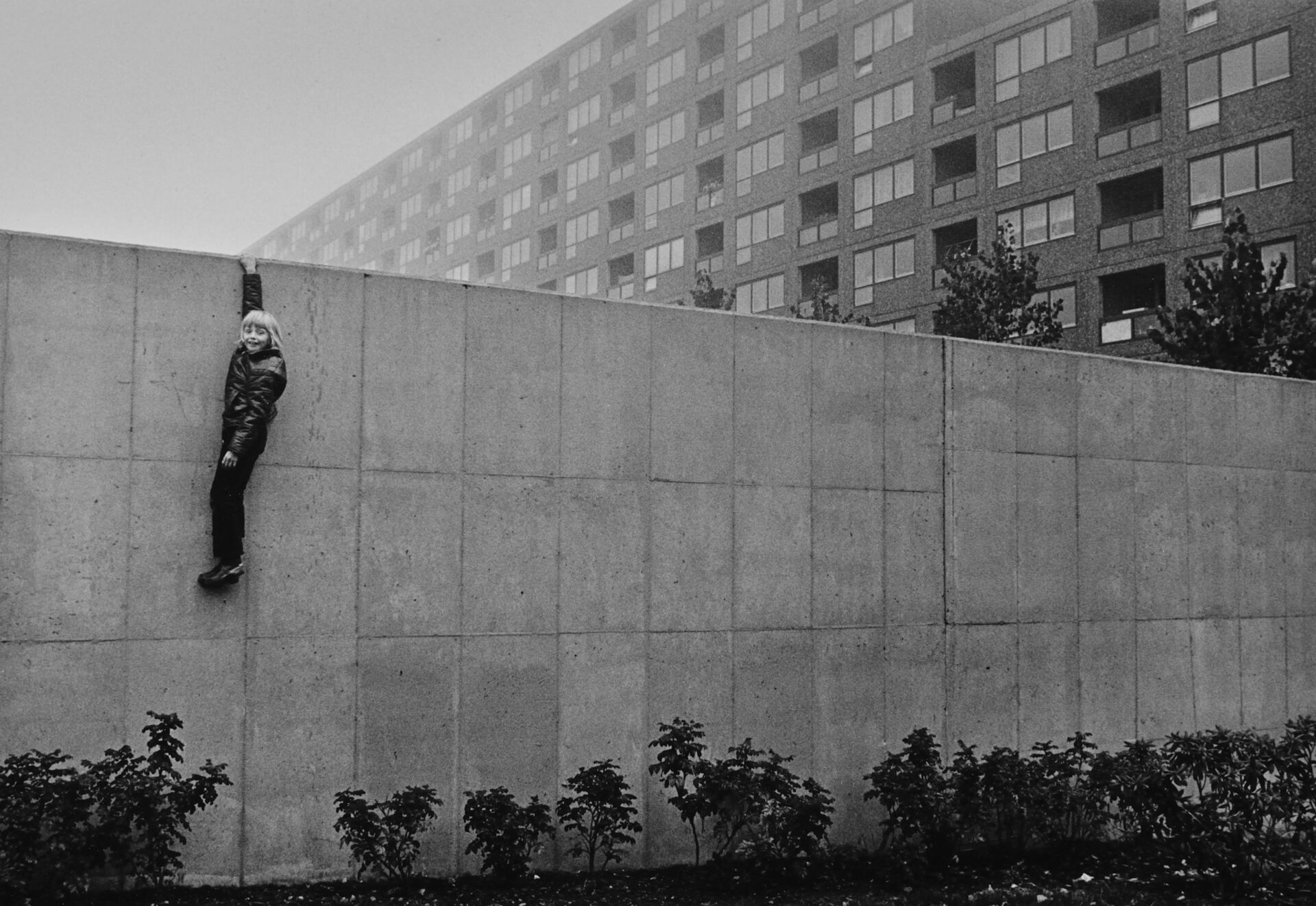 Svartvit bild föreställande en grå betonvägg och höghus i bakgrunden. En pojke hänger med ena handen på väggen.